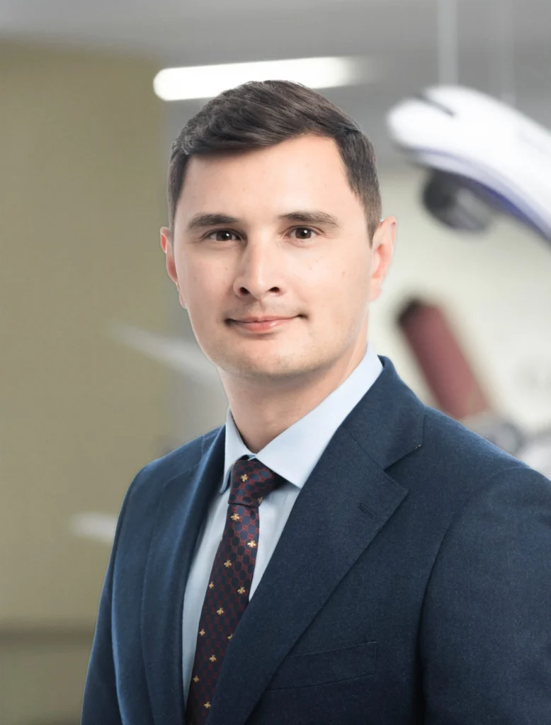 A person in a blue suit and tie stands in an office setting with blurred, modern equipment in the background.