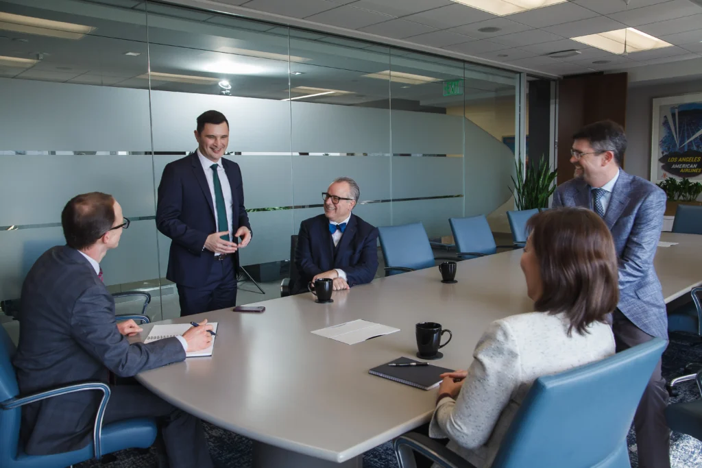 Group of professionals in business attire having a meeting in a modern conference room, with one person standing and presenting to colleagues seated around a large table.