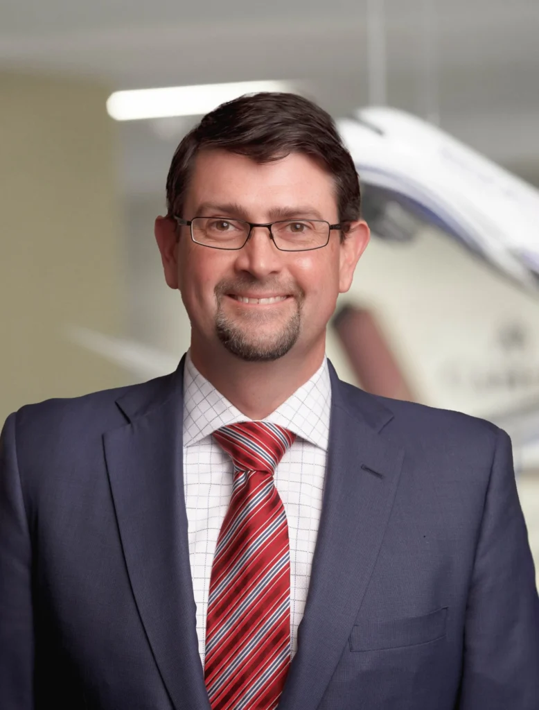 A man in a suit with glasses and a goatee smiles at the camera. He is wearing a red striped tie and a white checkered shirt. The background is a blurred office setting.