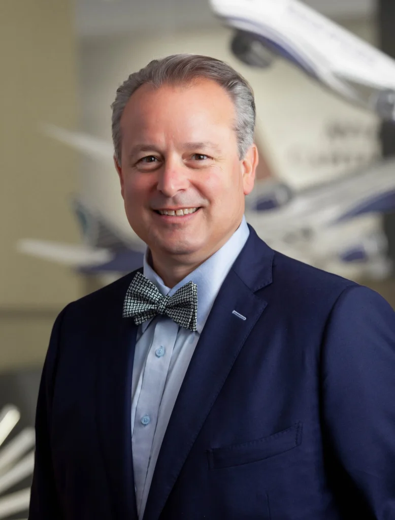 A man in a blue suit and bow tie smiles at the camera. He has short gray hair and is standing in front of a blurred background with model airplanes.