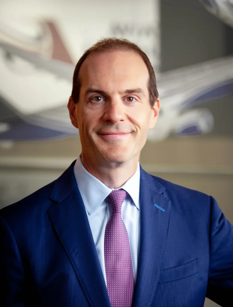 Professional man in a navy suit and pink tie, standing in an office environment with blurred airplane models in the background, highlighting aviation industry leadership and expertise.