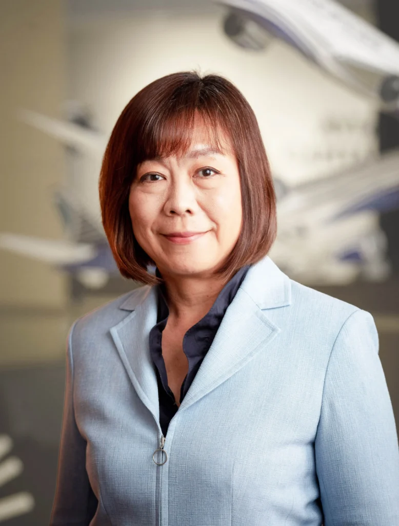 Professional woman in a light blue blazer, standing in an office environment with blurred airplane models in the background, representing aviation leadership and expertise.