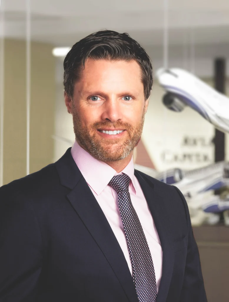 Professional man in a suit and tie smiling, standing in an office environment with blurred airplane models in the background, representing aviation industry expertise.