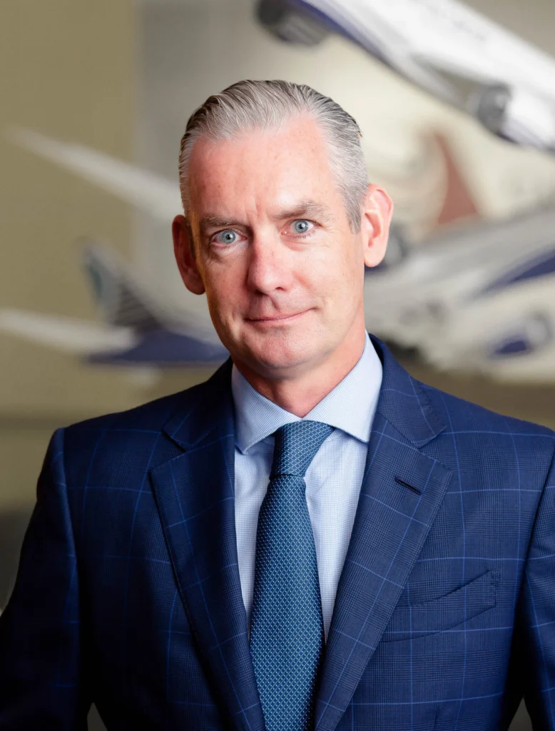 A man in a blue checkered suit and tie stands in front of an abstract background featuring blurred airplane models.