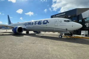 A United Airlines airplane is parked on the tarmac near a Boeing building under a partly cloudy sky. The aircraft is facing right, with visible landing gear and markings.