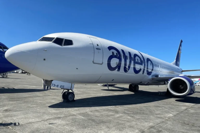 A white commercial airplane with avelo written on the side is parked on a tarmac under a clear blue sky. The aircraft is positioned diagonally, showing the front section and wing. Other planes are visible in the background.